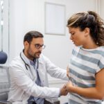 Patient parlant au médecin de sa douleur et de ses problèmes de santé lors d'une visite à l'hôpital Jeune femme se plaignant de douleurs au dos ou aux reins, assise sur la table d'examen dans le cabinet du médecin. meilleurs médecins spécialistes de l'appareil digestif
