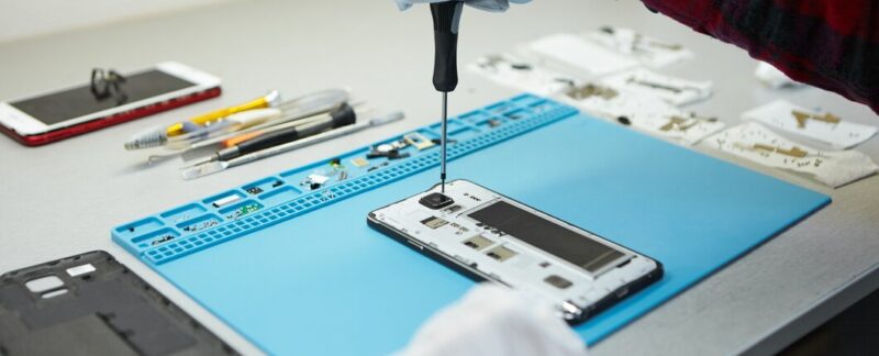 Technicien portant des gants antistatiques utilisant un tournevis pour démonter un téléphone portable endommagé, allant réparer la carte mère, assis à son poste de travail dans le laboratoire avec l'équipement nécessaire. Les meilleurs centres de réparation de téléphones portables en France