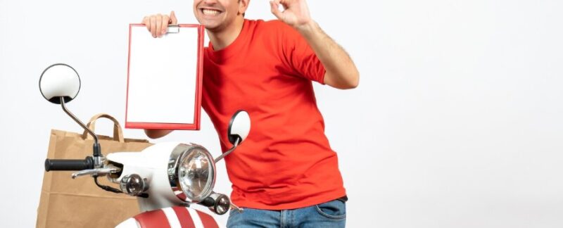 Vue de dessus d'un jeune livreur souriant, vêtu d'un uniforme rouge, debout près d'une moto. Top 10 des services gratuits en France